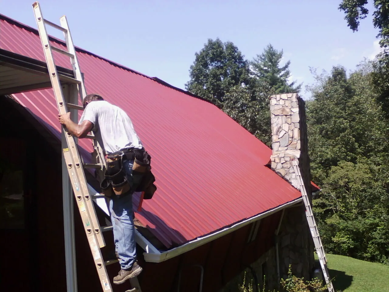 Roofer on a ladder beside a red metal roof and stone chimney — Soffit & Fascia Repair by Roofing Brothers in Long Branch